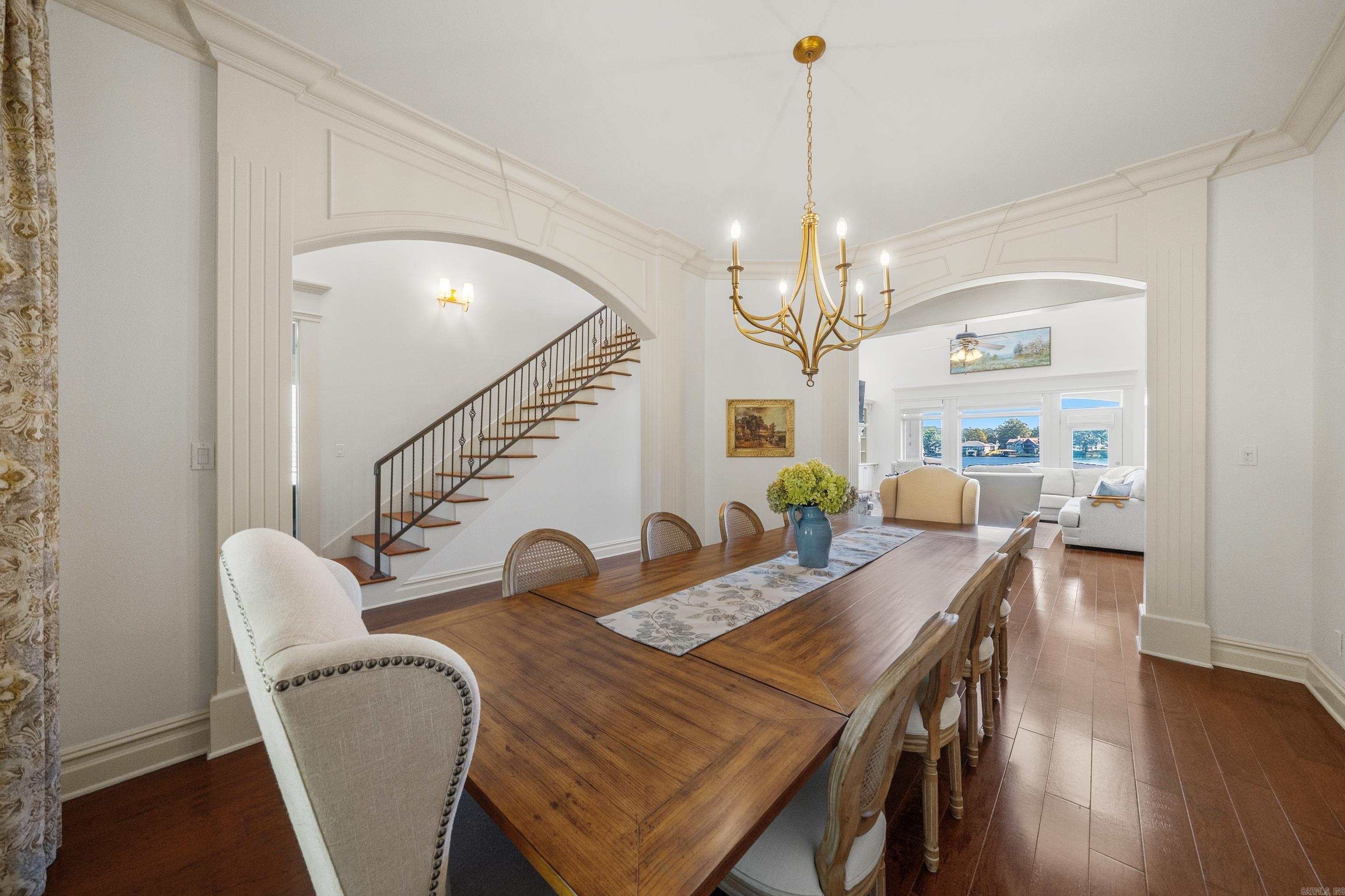 Dining room with a long wooden table and brass chandelier, opening to a sitting room with water views.