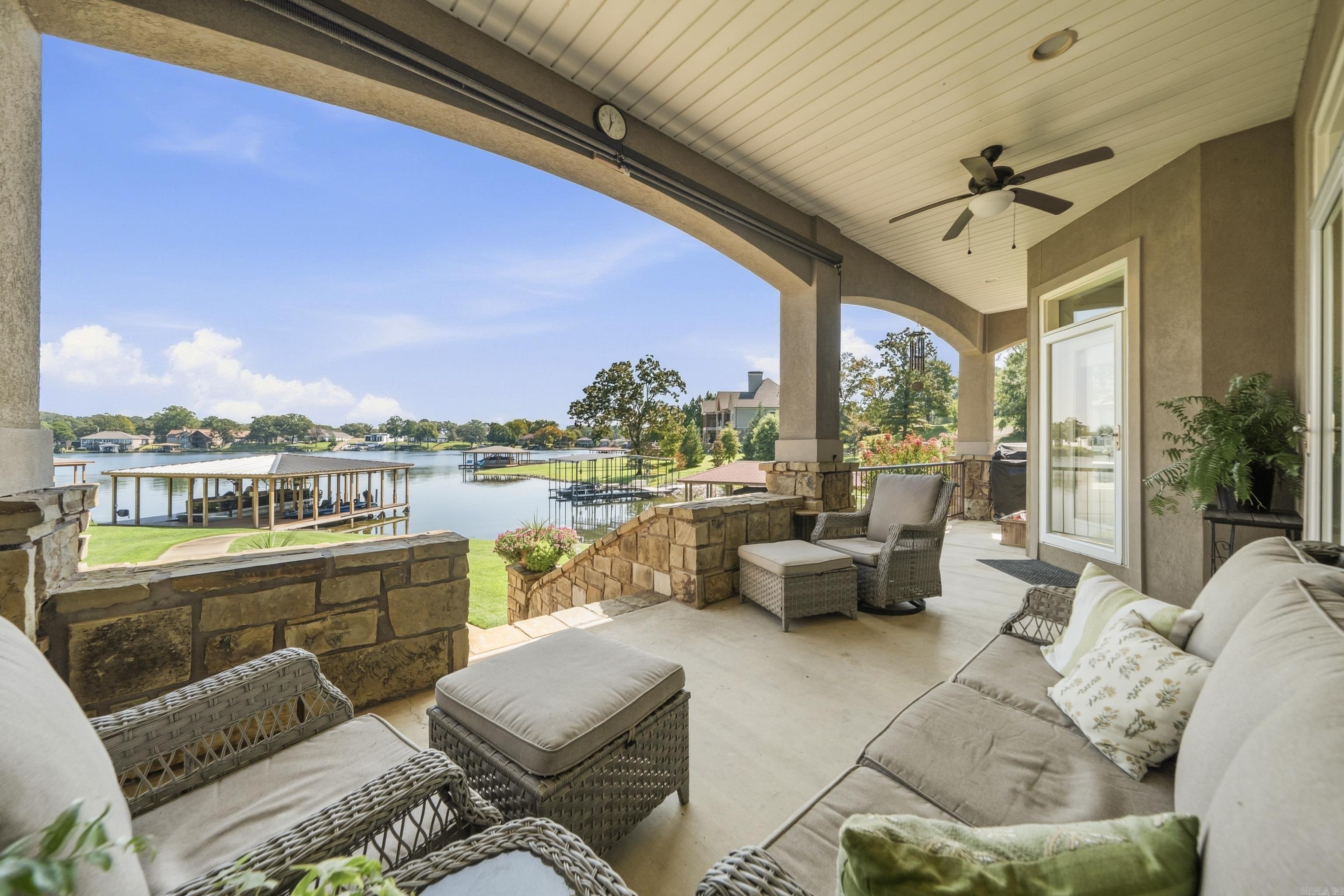 Furnished covered porch overlooking a lake, with wicker seating, stone columns and boat docks.