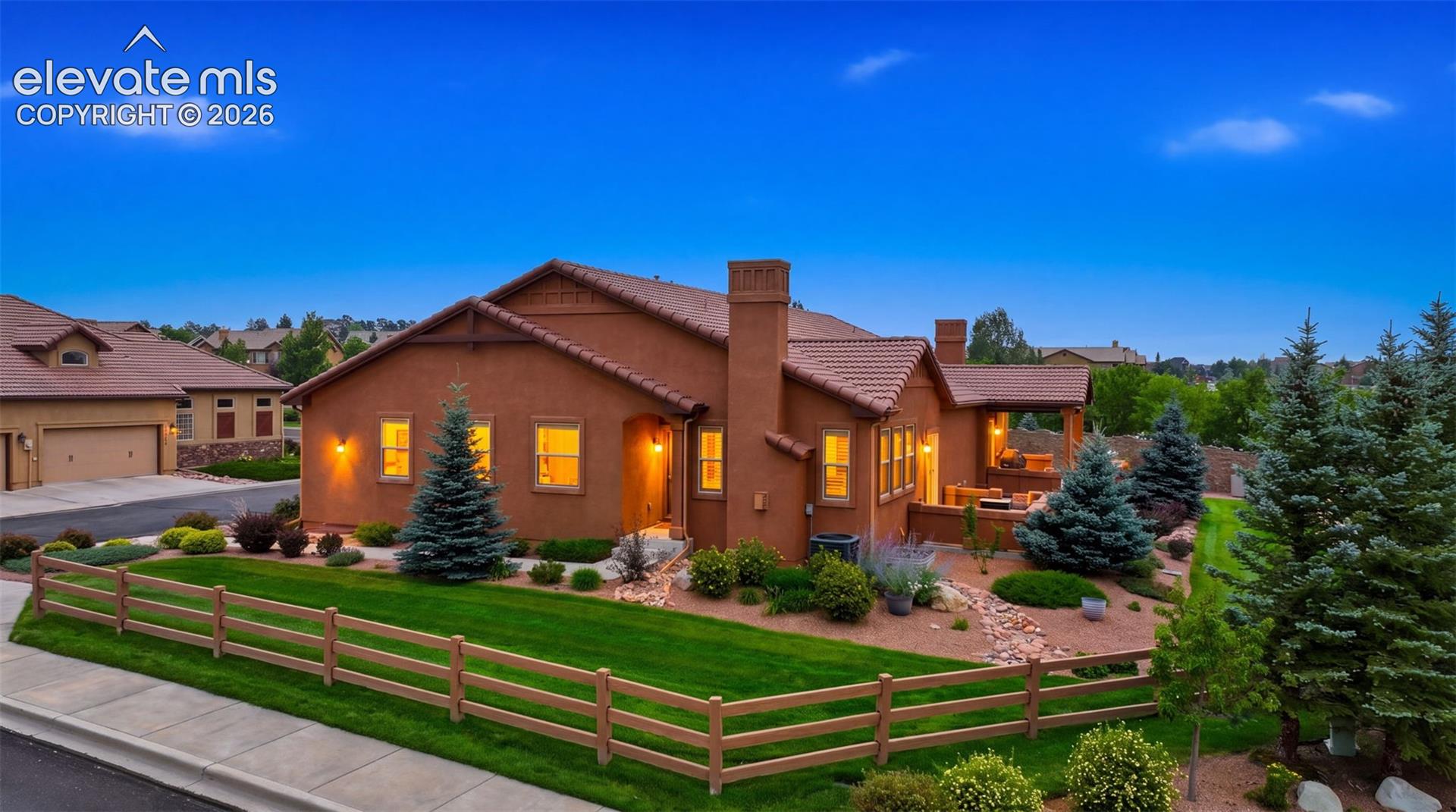 Reddish stucco home with a tile roof, fenced lawn, and covered patio at dusk.