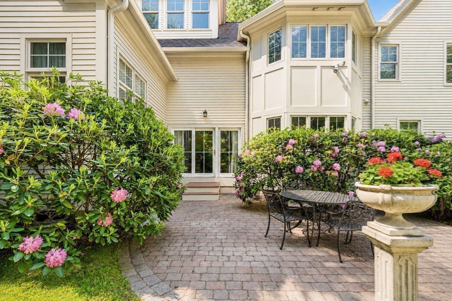Rear patio with brick pavers, outdoor dining set, flowering shrubs, and a stone fountain.