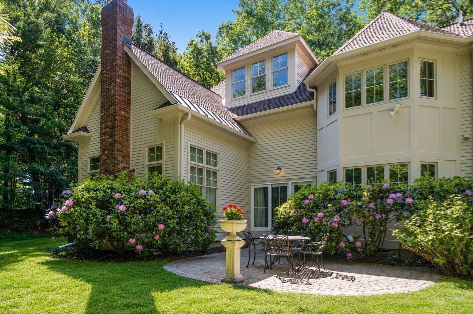 Home’s rear exterior with a brick chimney, flowering shrubs, and a stone patio with outdoor dining furniture.
