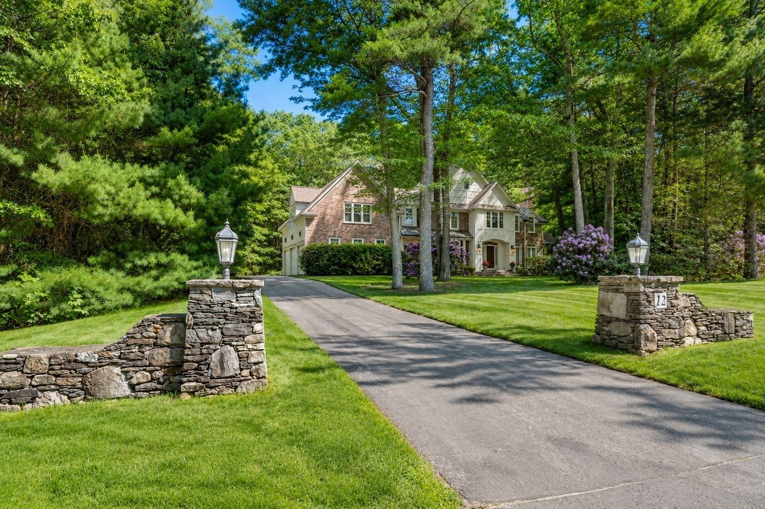 Large stone-and-siding home beyond a paved driveway, framed by mature trees and stone entrance walls.