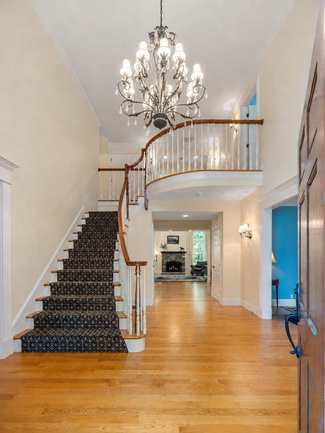 Two-story foyer with a sweeping carpeted staircase, curved balcony, chandelier, and hardwood floors.