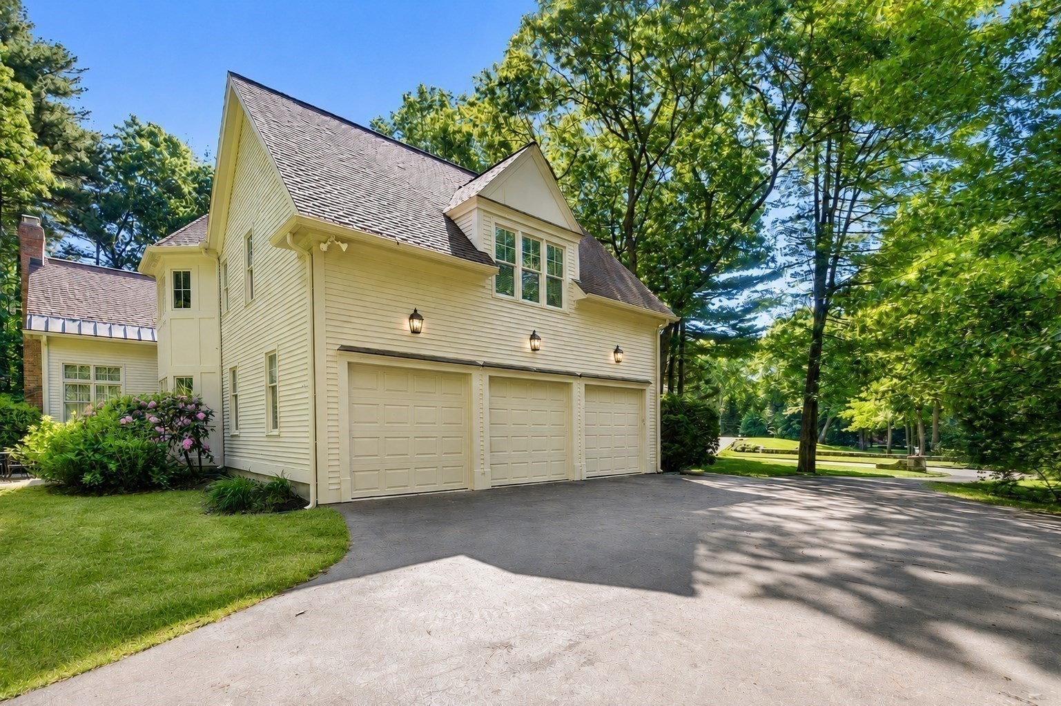 Light-colored two-story house with three garage doors, a gabled roof, and mature trees.