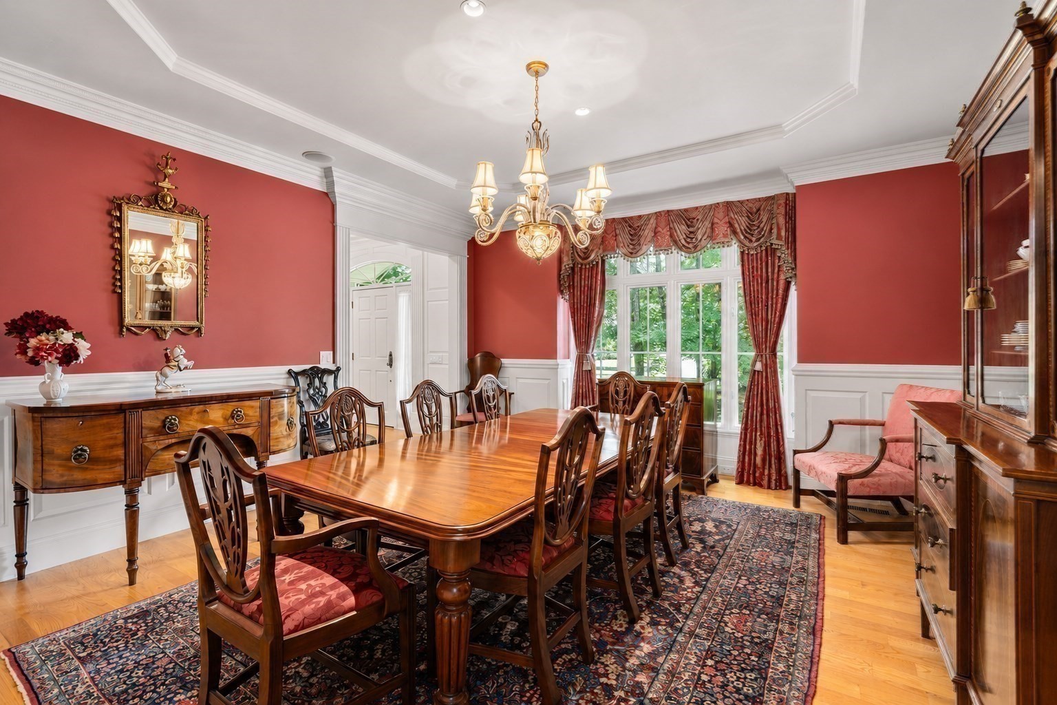 Dining room with a polished wood table, carved chairs, red walls, and tall curtained windows.