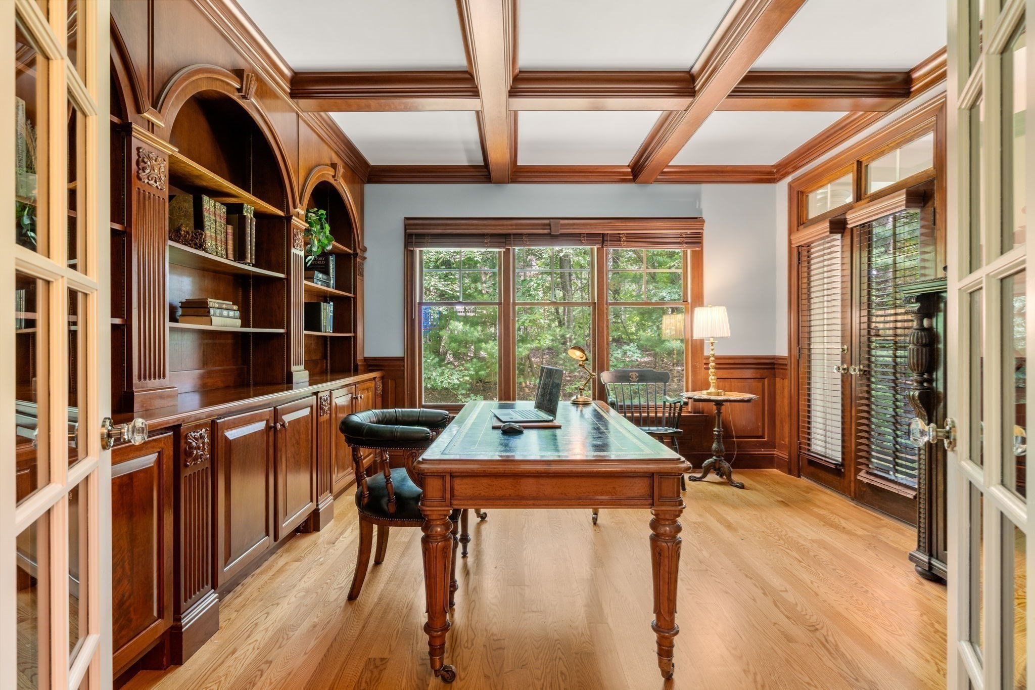 Home office with built-in mahogany bookcases, a wooden desk, coffered ceiling, and large garden-facing windows.
