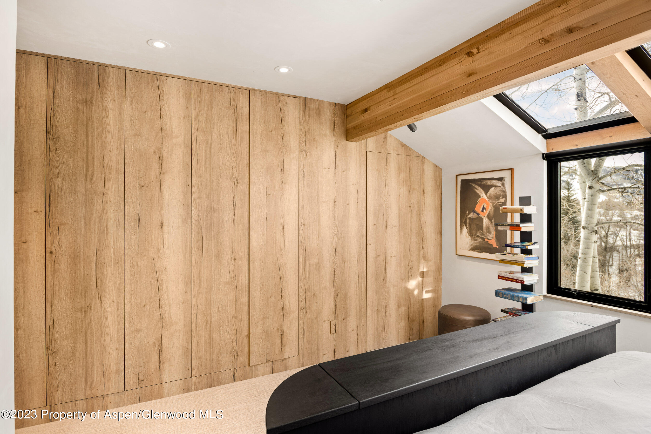 Bedroom with a tall wood-paneled wall, dark bed frame, skylight, and window overlooking bare trees.