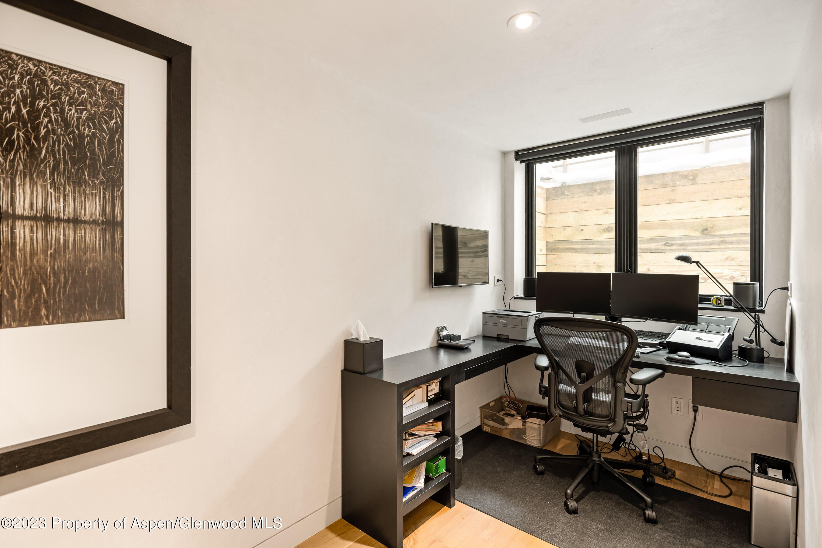 Home office with a dark built-in desk, dual monitors, and a large window overlooking a wood fence.