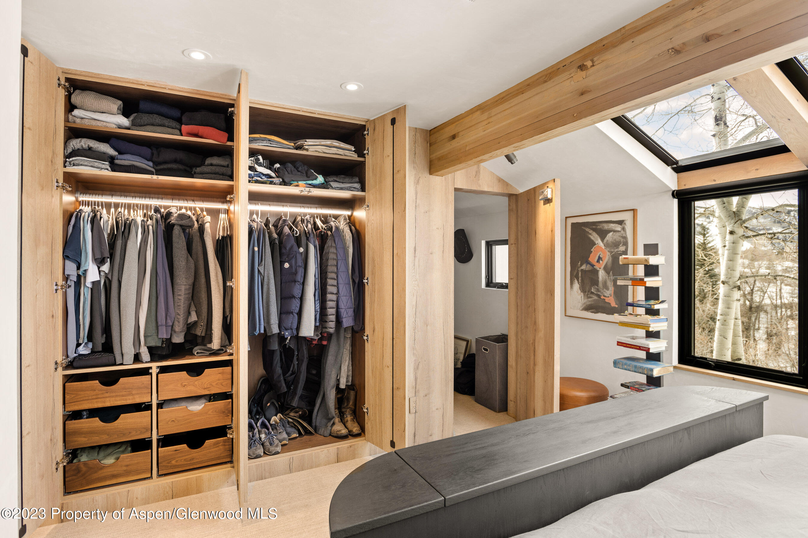 Bedroom with illuminated built-in closets, organized clothing, and a large window overlooking aspen trees.