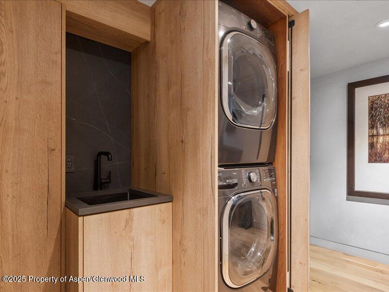 Laundry area with stacked washer and dryer beside a dark utility sink in warm wood cabinetry.