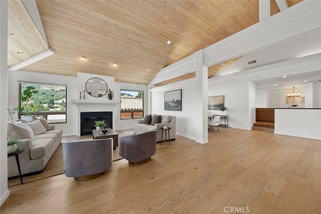 Living room with vaulted wood ceiling, white beams, fireplace, and windows overlooking green hills.