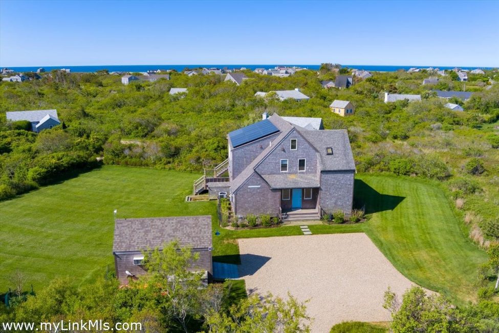 Aerial view of a shingle-sided home with a detached outbuilding, gravel driveway, and ocean beyond.
