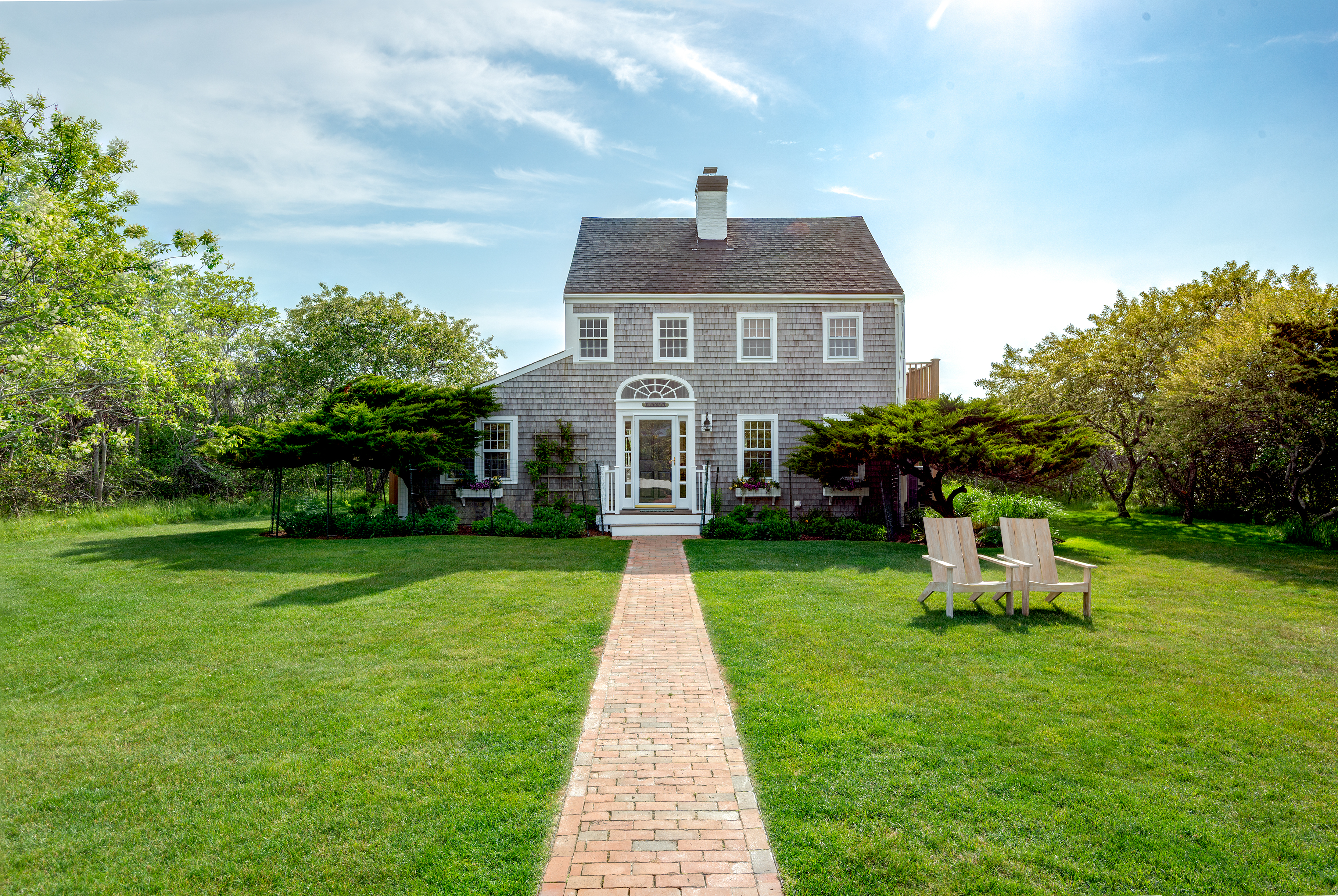 Gray-shingled two-story home beyond a brick walkway, with a wide lawn and two Adirondack chairs.