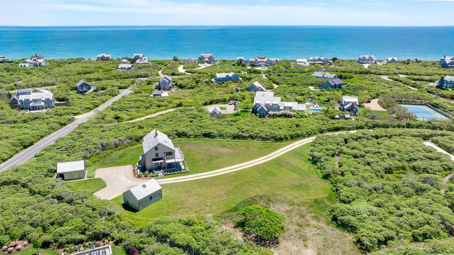 Aerial view of a shingled coastal home, surrounding scrubland, neighboring houses, and ocean.