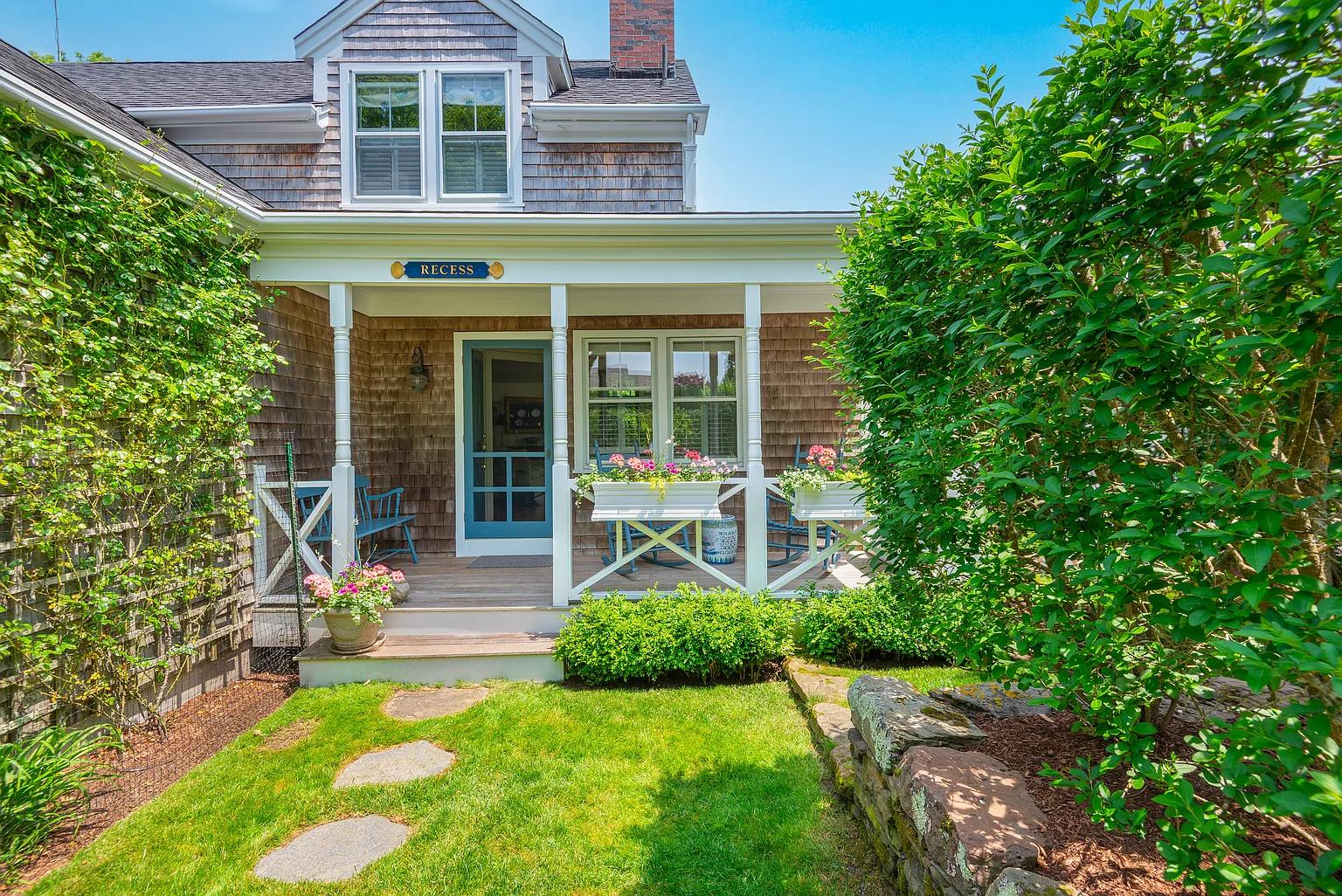 Shingle-style home with a covered porch, blue door, flower boxes, and leafy garden.