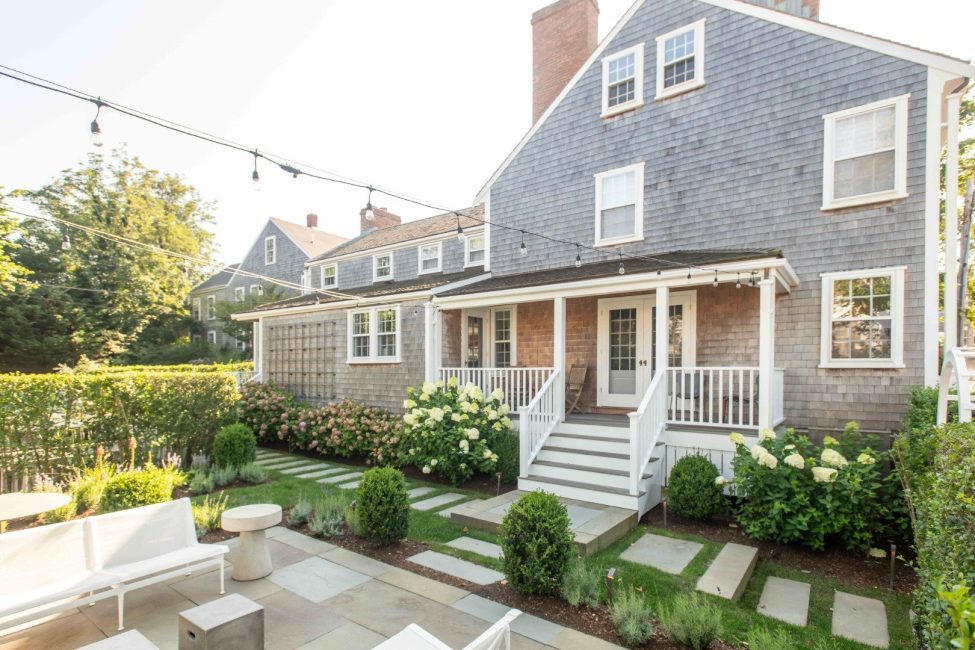 Gray-shingled house with a white-railed porch, landscaped garden, and patio seating.