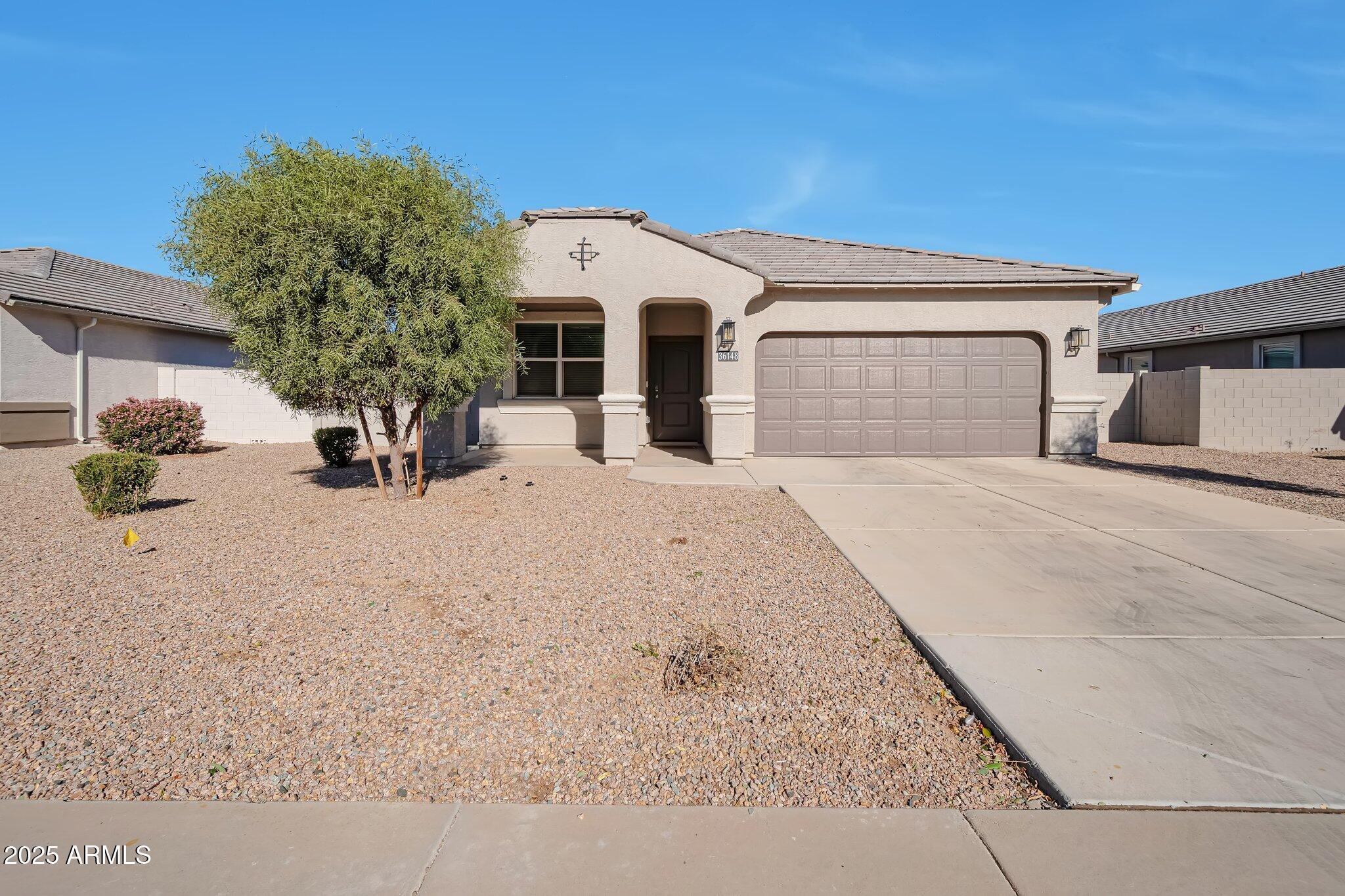 Single-story stucco home with a two-car garage, gravel yard, and leafy front-yard tree.