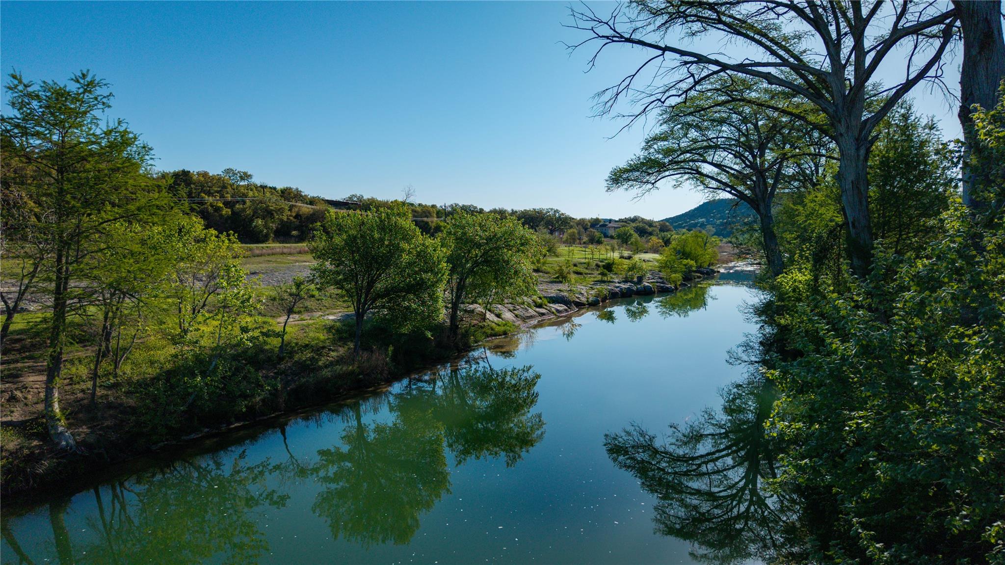 Calm creek reflecting leafy trees, with rocky banks and wooded hills beyond.