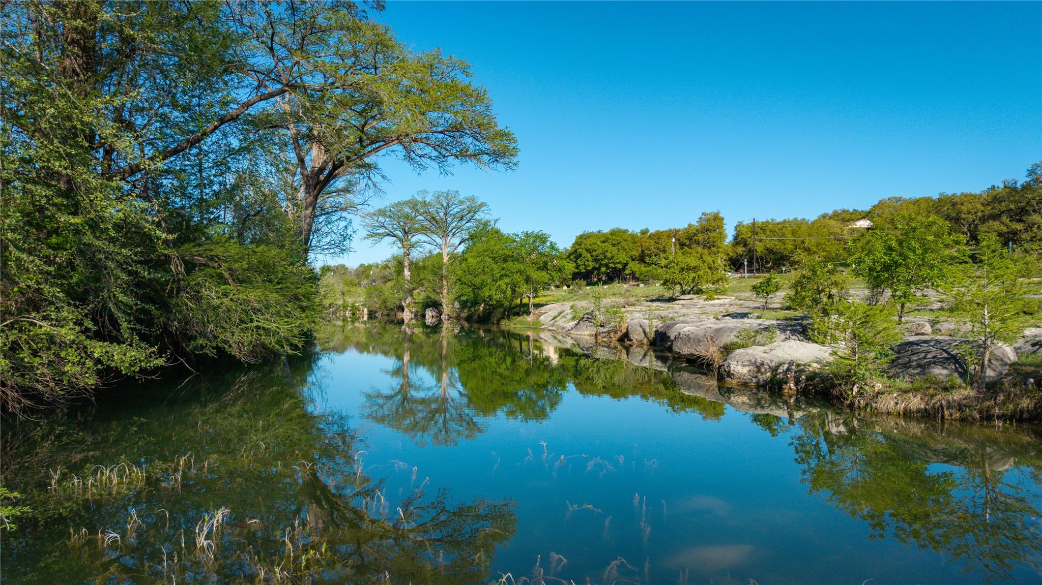 Rocky creek lined with leafy trees, reflected in calm water beneath a clear blue sky.