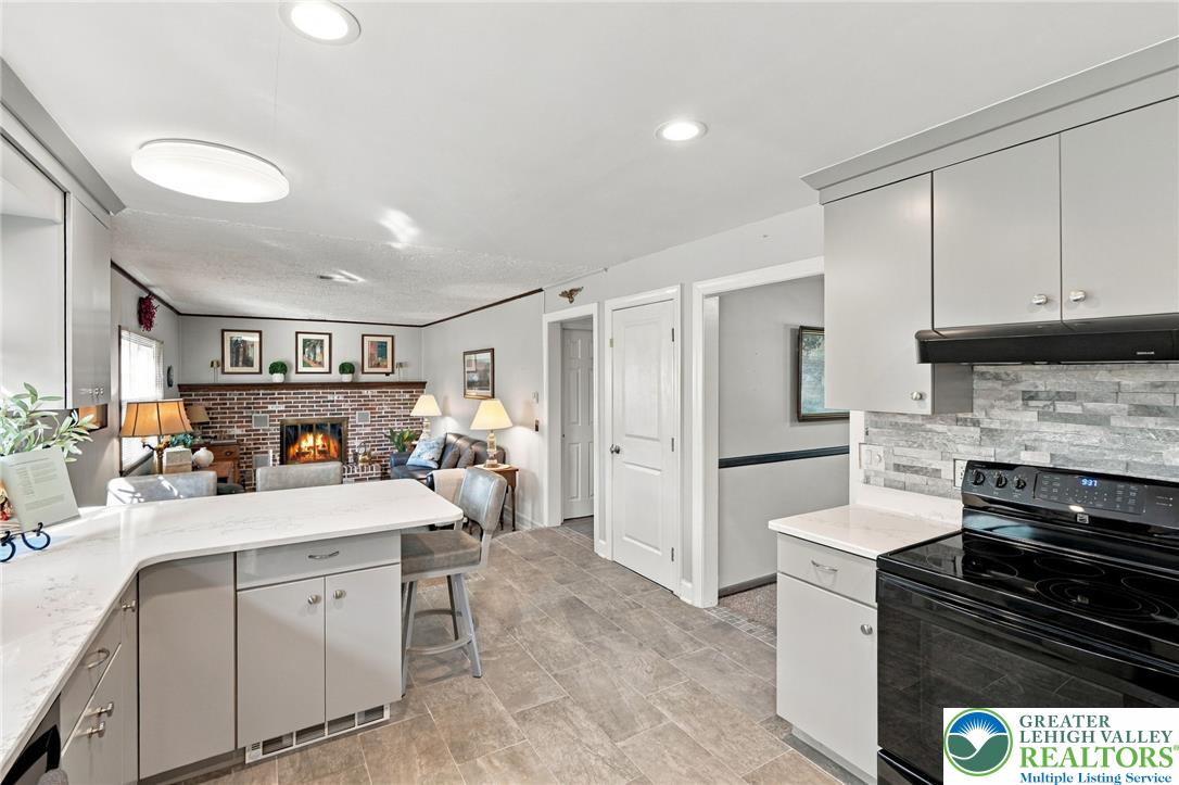 Kitchen with white cabinets and gray tile backsplash, open to a living room with a brick fireplace.