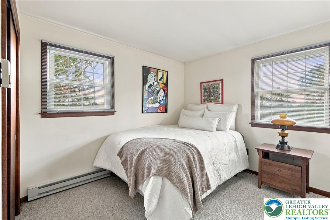 Bedroom with a white bed, taupe throw, two bright windows, and framed artwork.