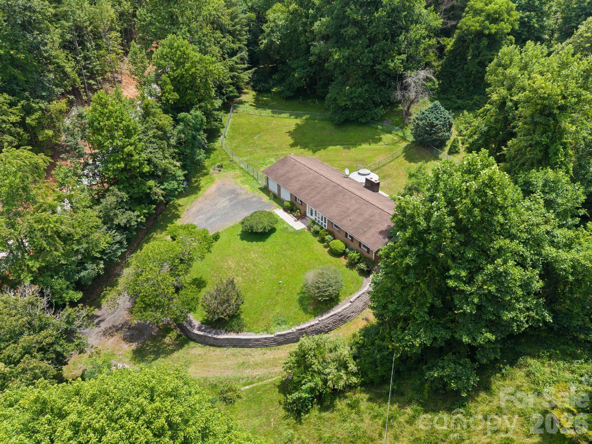 Aerial view of a brick house with a fenced lawn, gravel driveway, and curved stone retaining wall.