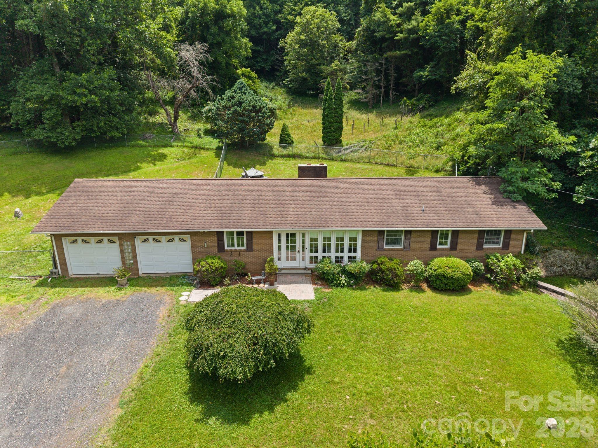 Aerial view of a brick ranch home with two garage doors, a gravel driveway, and a wooded backyard.