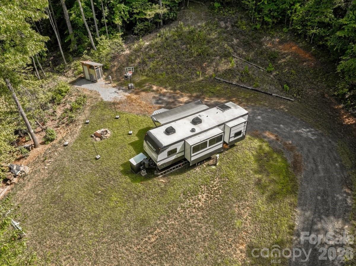 A travel trailer in a wooded clearing with a shed, basketball hoop, and gravel drive.