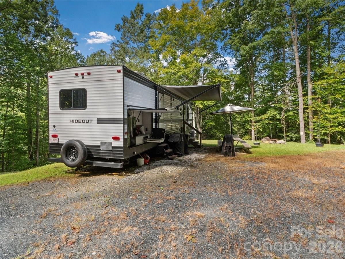 Travel trailer in a wooded gravel clearing with an extended awning and outdoor seating.