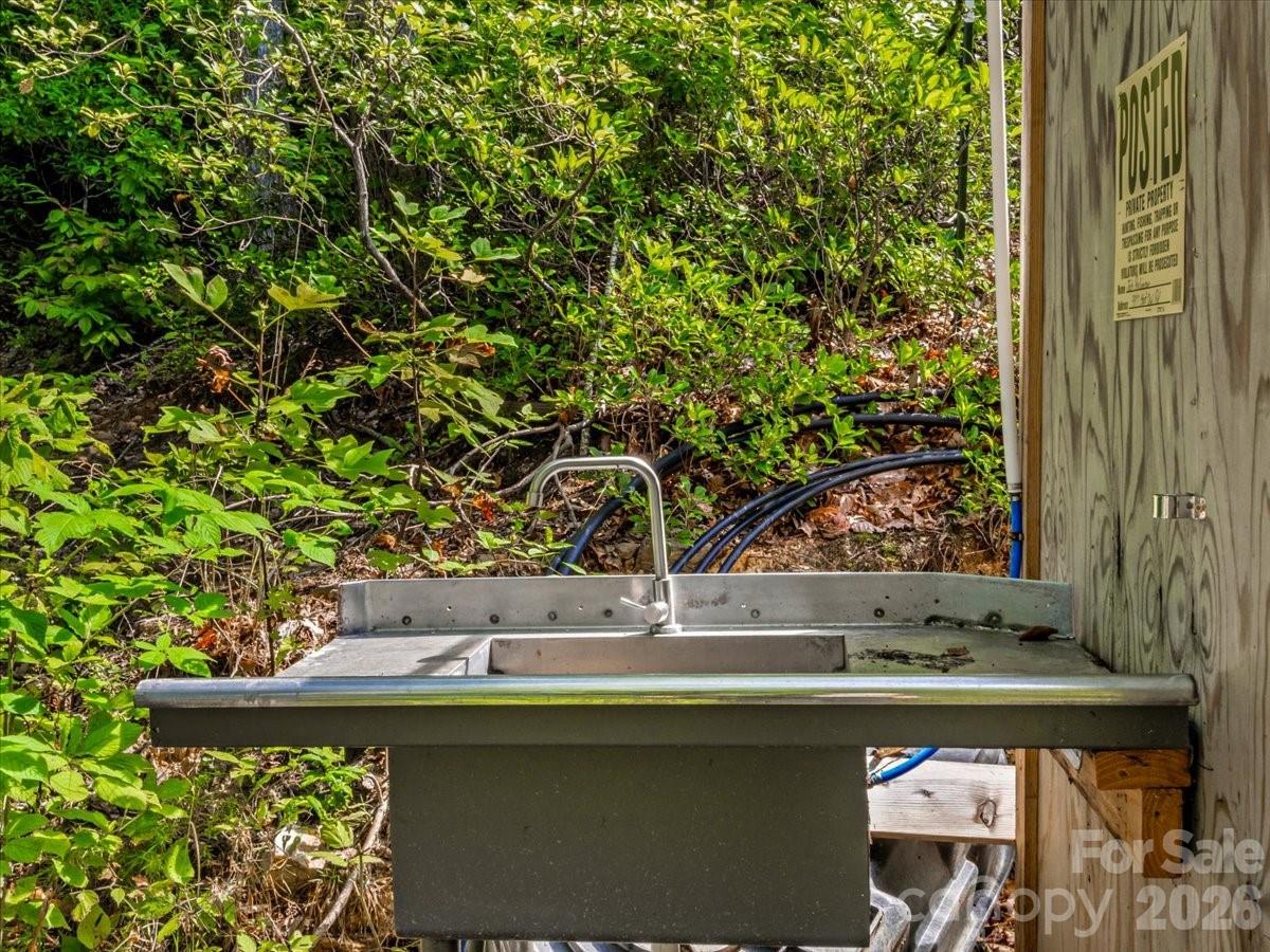 Outdoor metal utility sink beside a wooden wall, with hoses and dense woodland behind it.