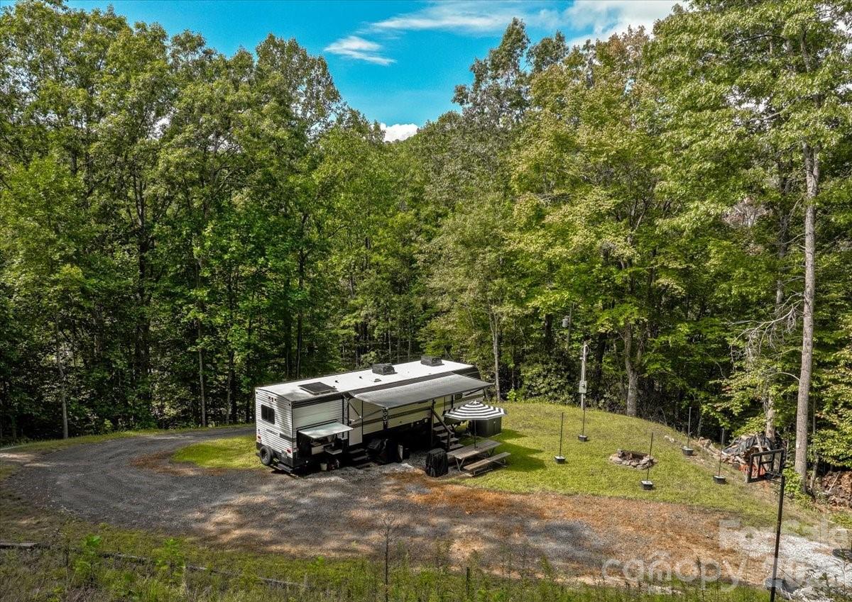 Aerial view of a travel trailer in a wooded clearing beside a gravel driveway and fire pit.