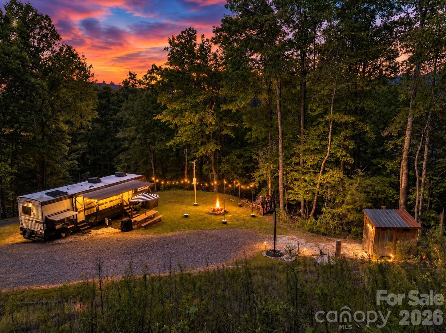Wooded campsite with a travel trailer, glowing string lights, and a fire pit at sunset.