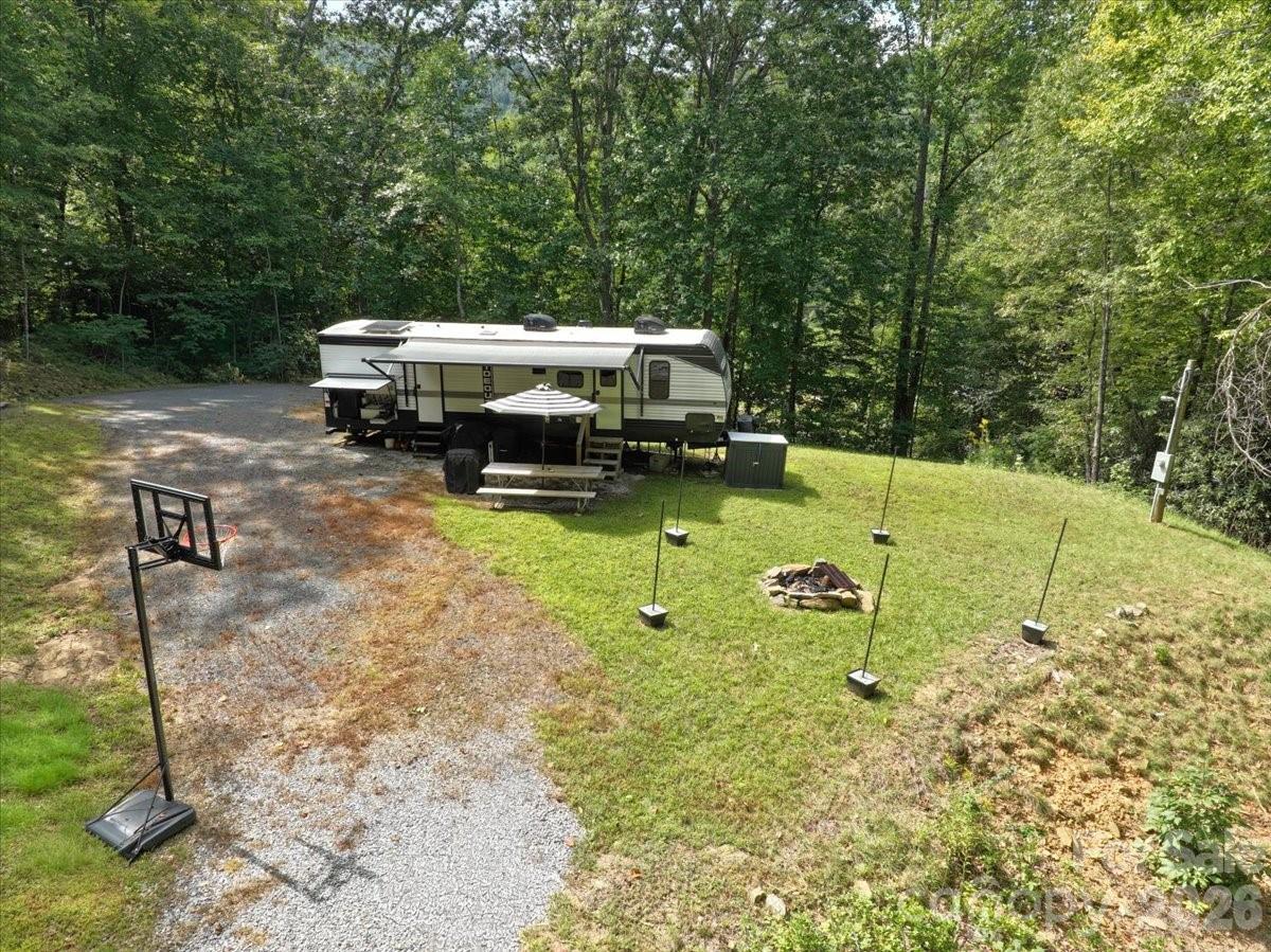 Travel trailer in a wooded clearing beside a gravel driveway, basketball hoop, and fire pit.