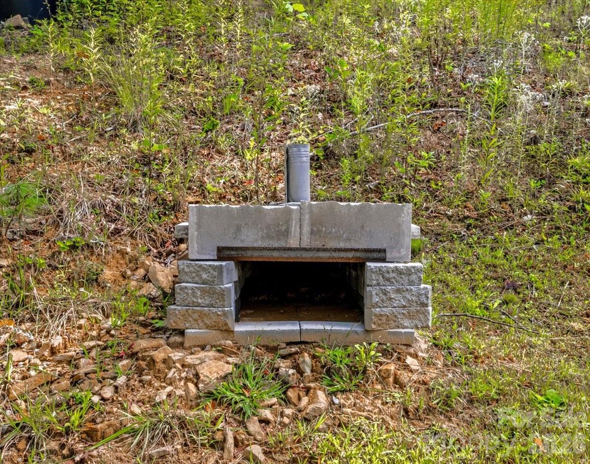 Outdoor masonry fireplace with an open firebox and short metal flue on a wooded slope.