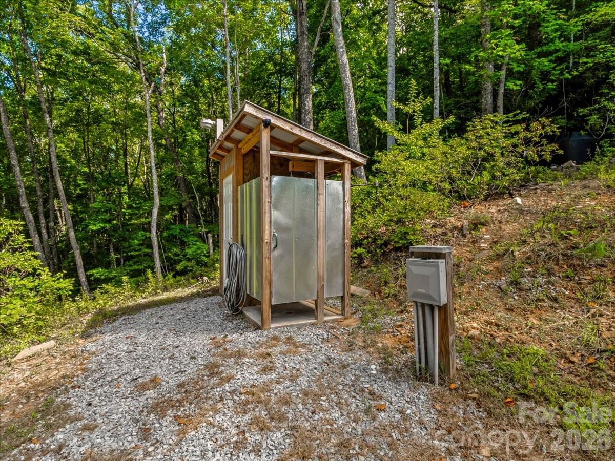 Wooden utility enclosure with metal panels and a coiled hose in a wooded gravel clearing.