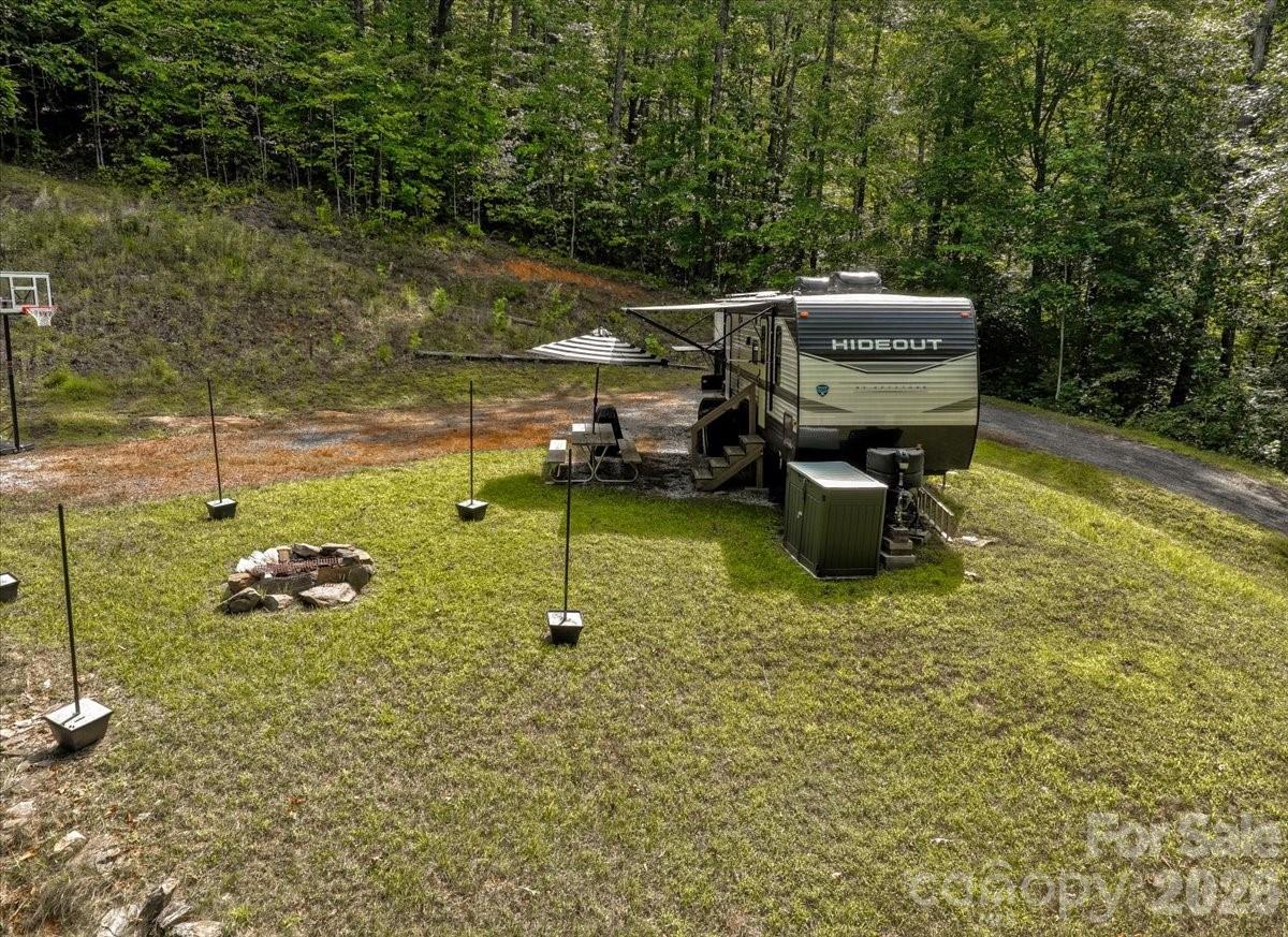 Grassy wooded clearing with a Hideout travel trailer, picnic table, basketball hoop, and fire pit.