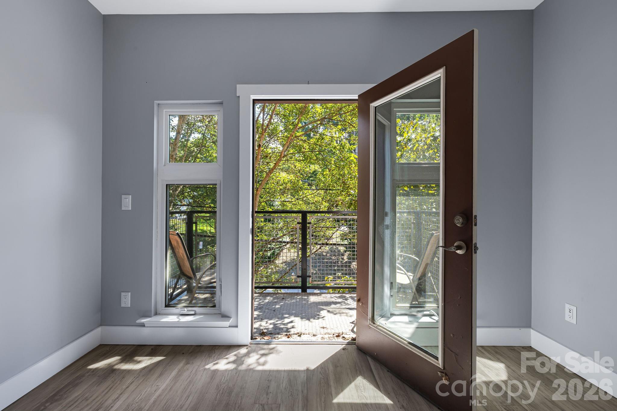 Open glass door from a vacant room onto a tree-shaded balcony, with a narrow window beside it.