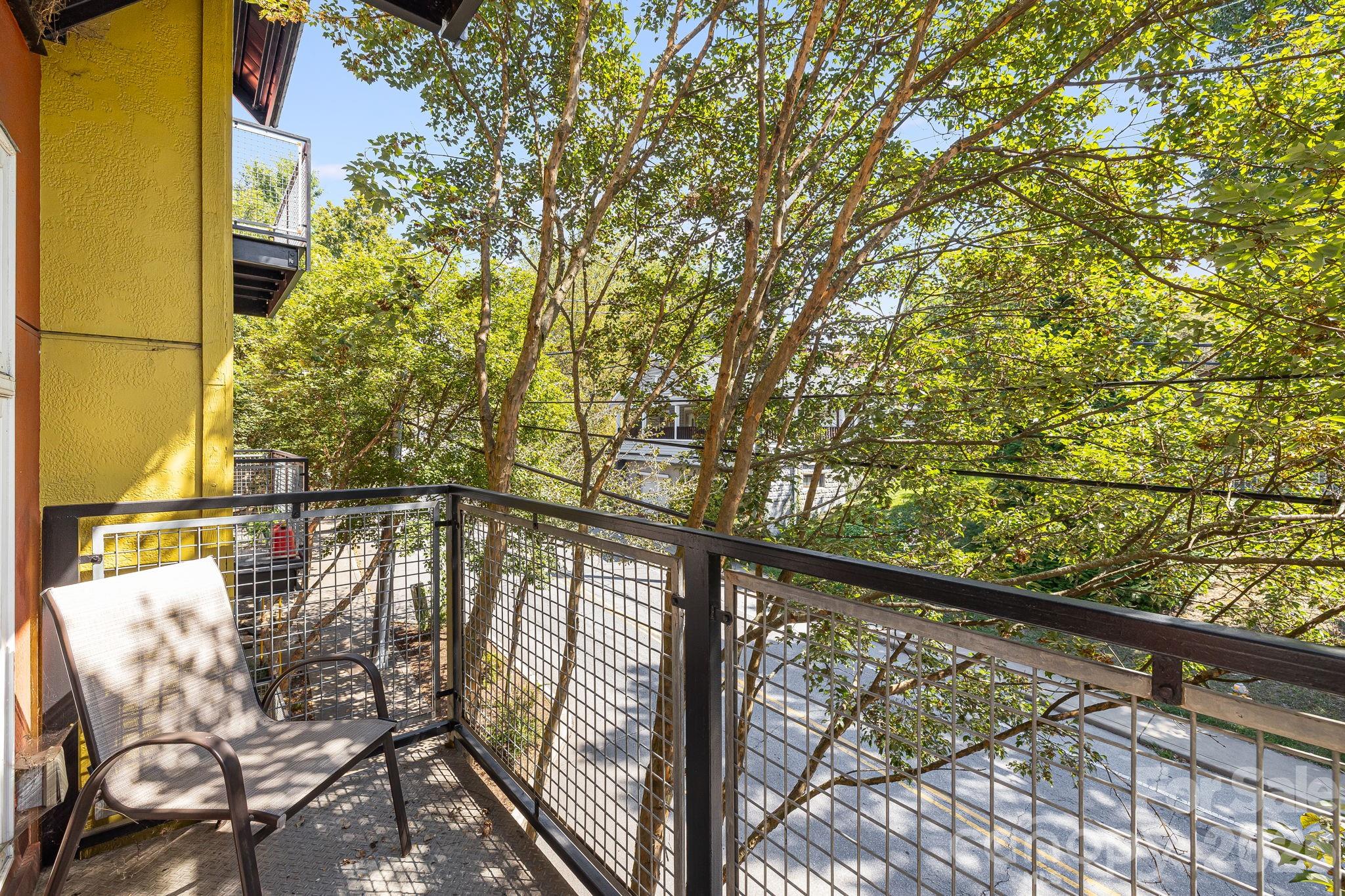 Apartment balcony with a mesh railing, chair, and leafy trees overlooking the street.