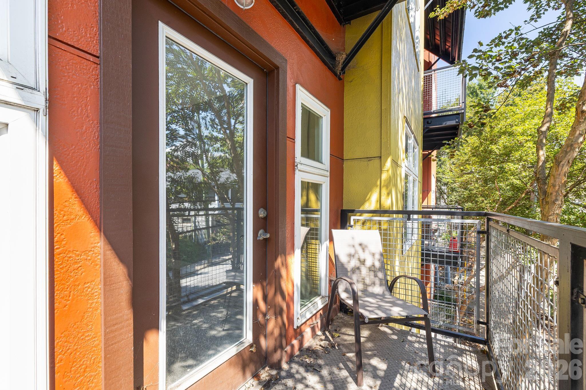 Condominium balcony with a metal railing, chair, and colorful exterior walls beside leafy trees.