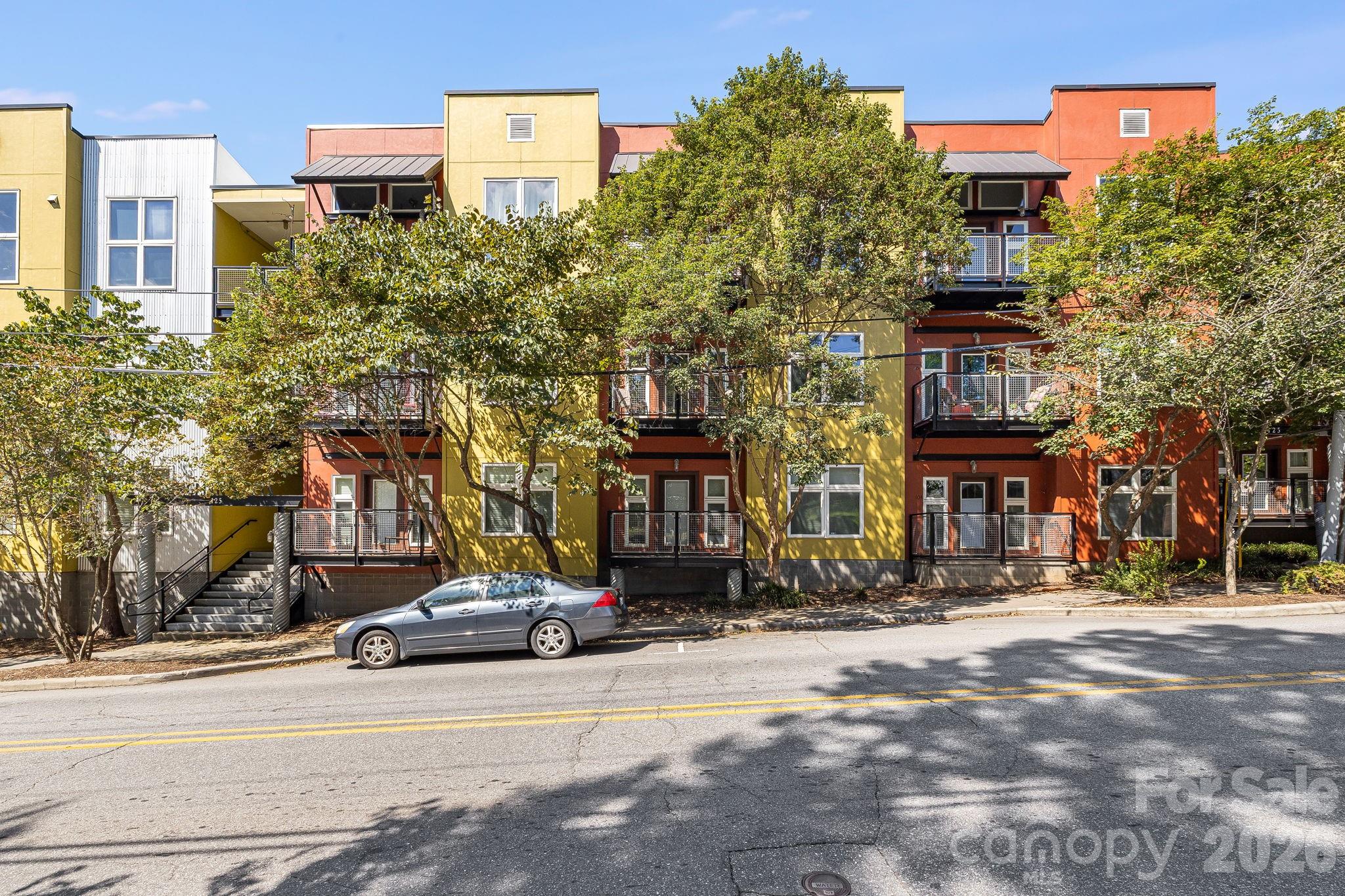 Colorful condominium building with metal balconies and leafy trees along the street.