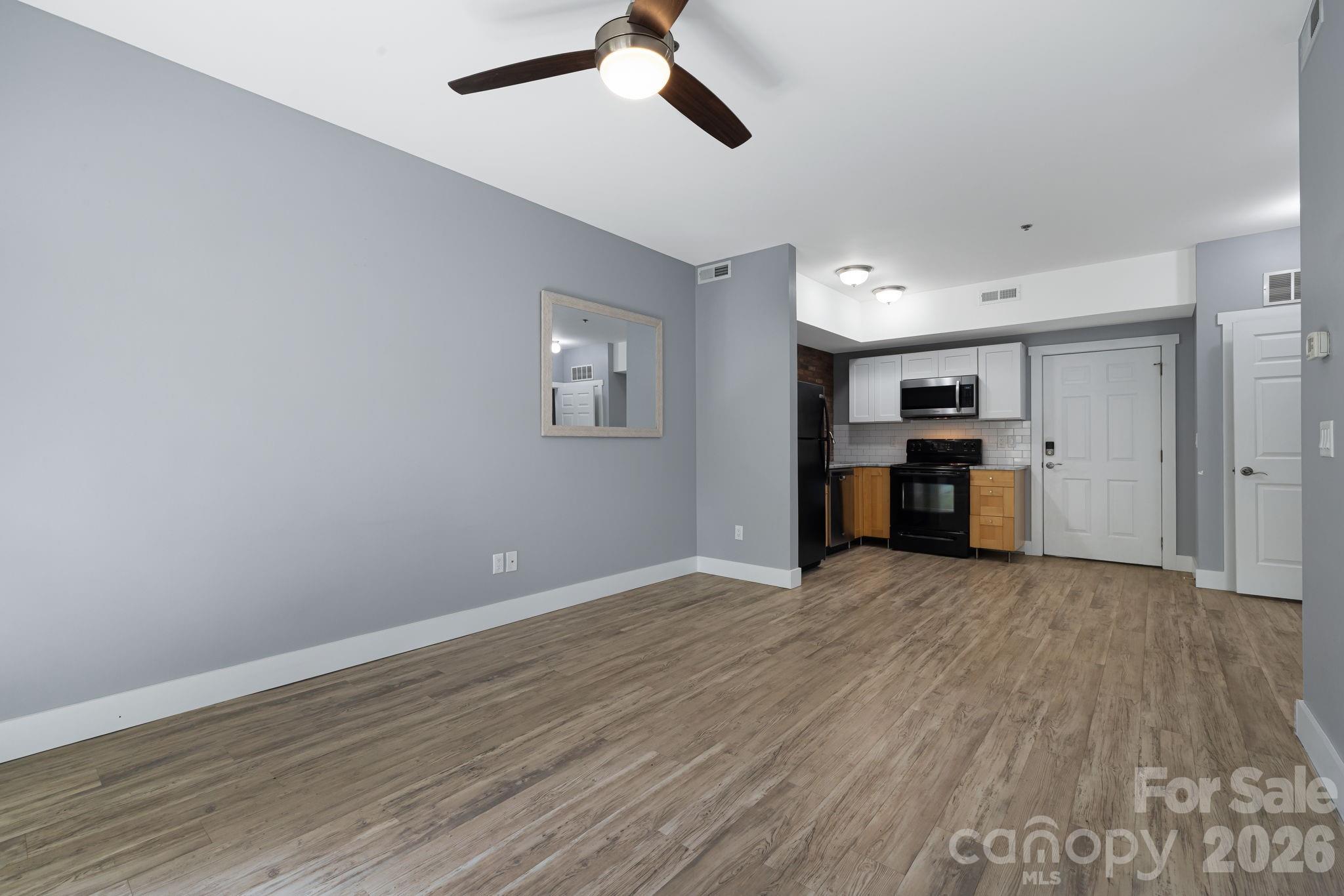 Vacant condo living area with wood-look floors, gray walls, and an open kitchen with black appliances.
