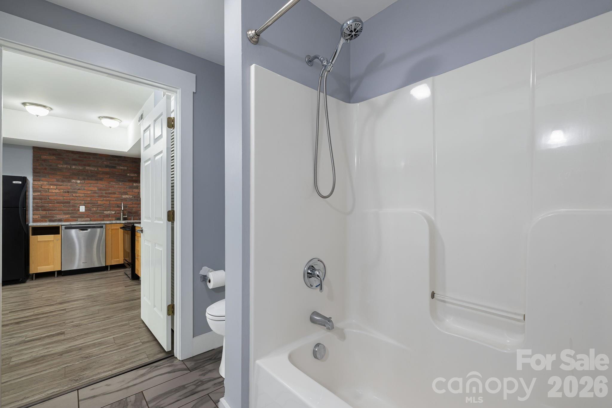 Bathroom tub and shower with a handheld showerhead; the open doorway reveals a kitchen with a brick backsplash.