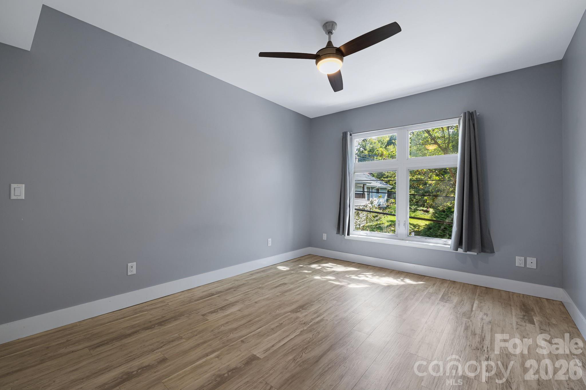 Empty room with gray walls, wood-look floors, a curtained window, and a ceiling fan.