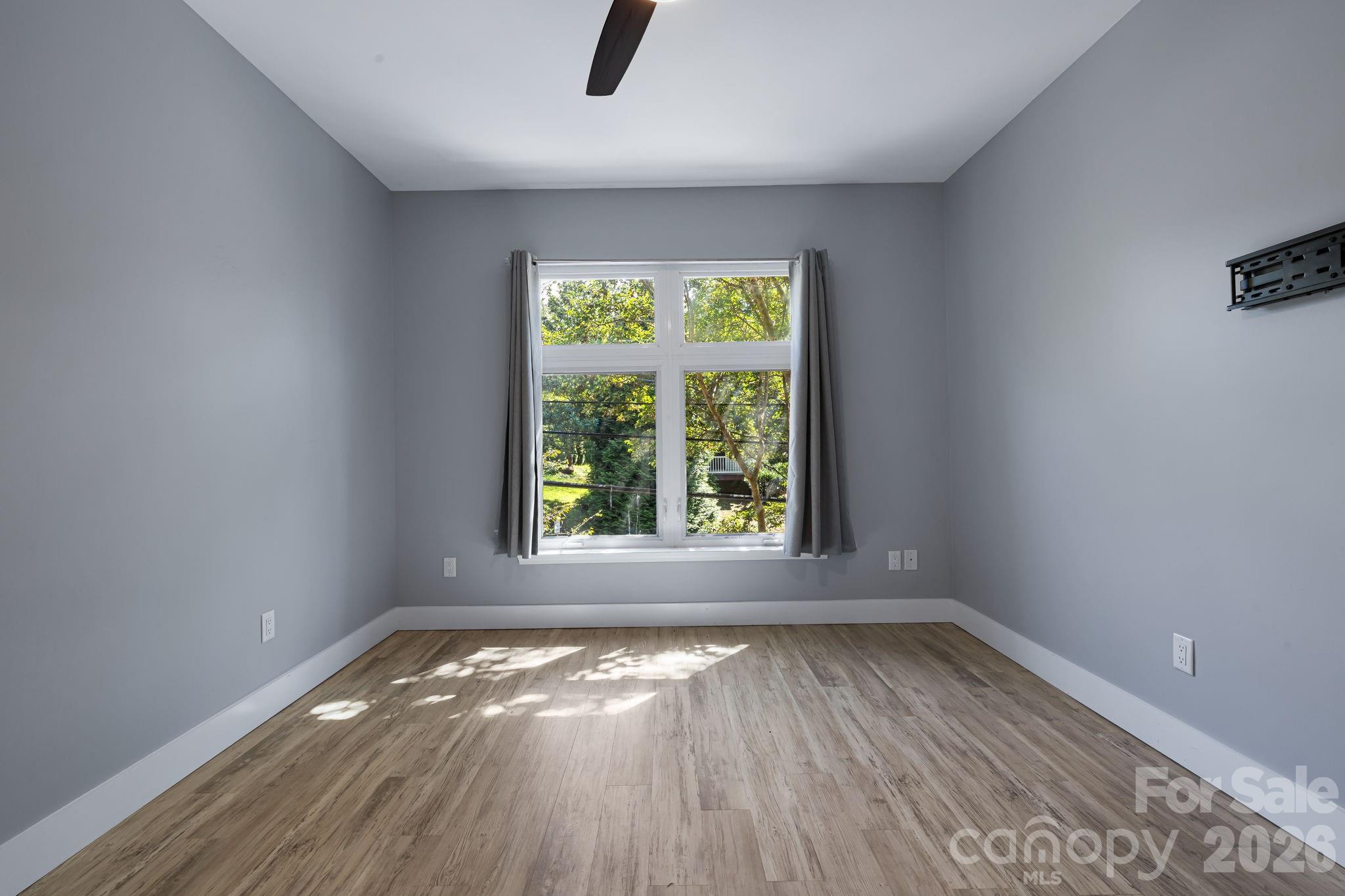 Vacant bedroom with wood-look flooring, a large curtained window, and a wall-mounted TV bracket.