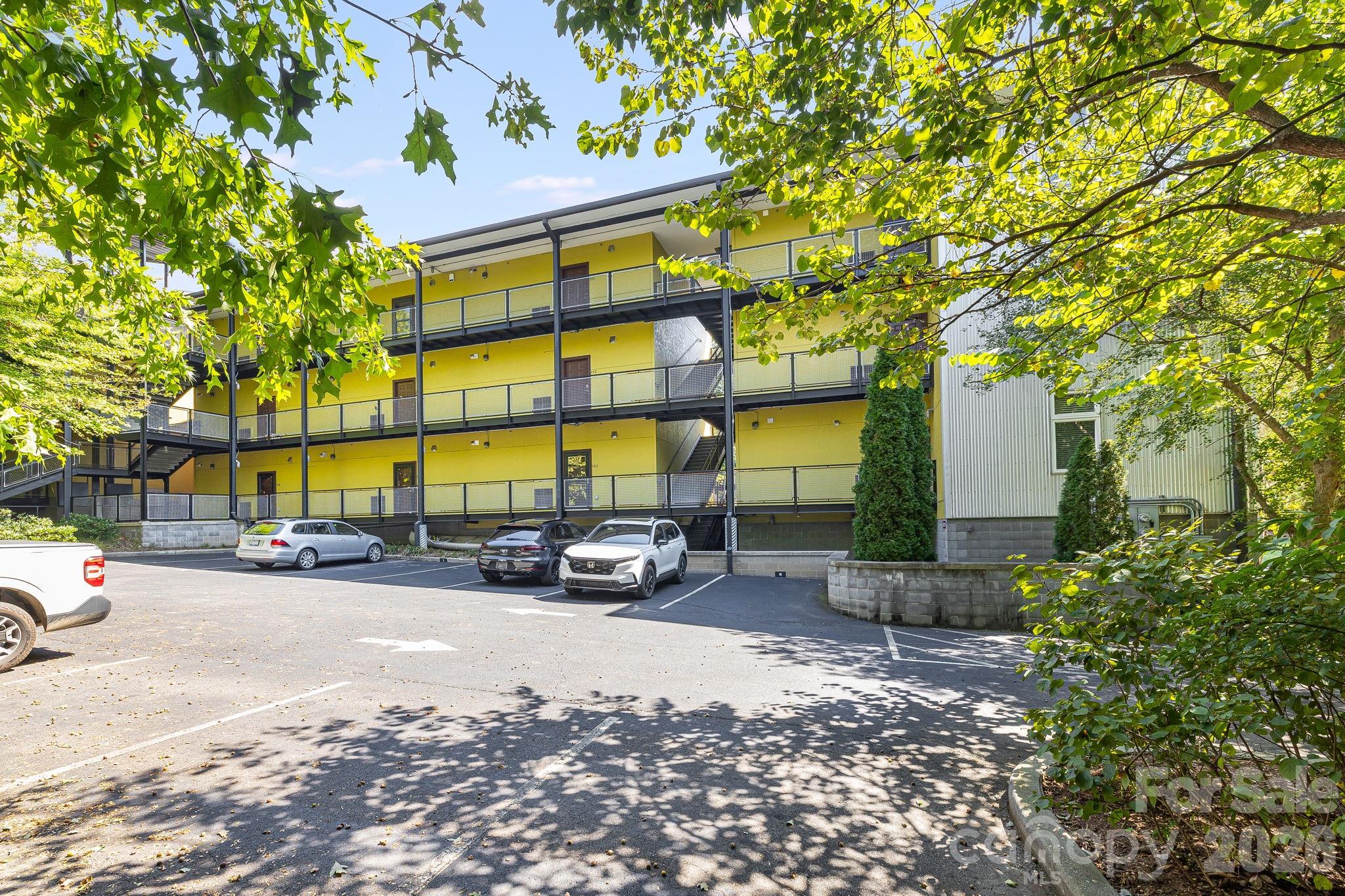 Three-story yellow apartment building with black-railed balconies, exterior stairs, and a parking lot.