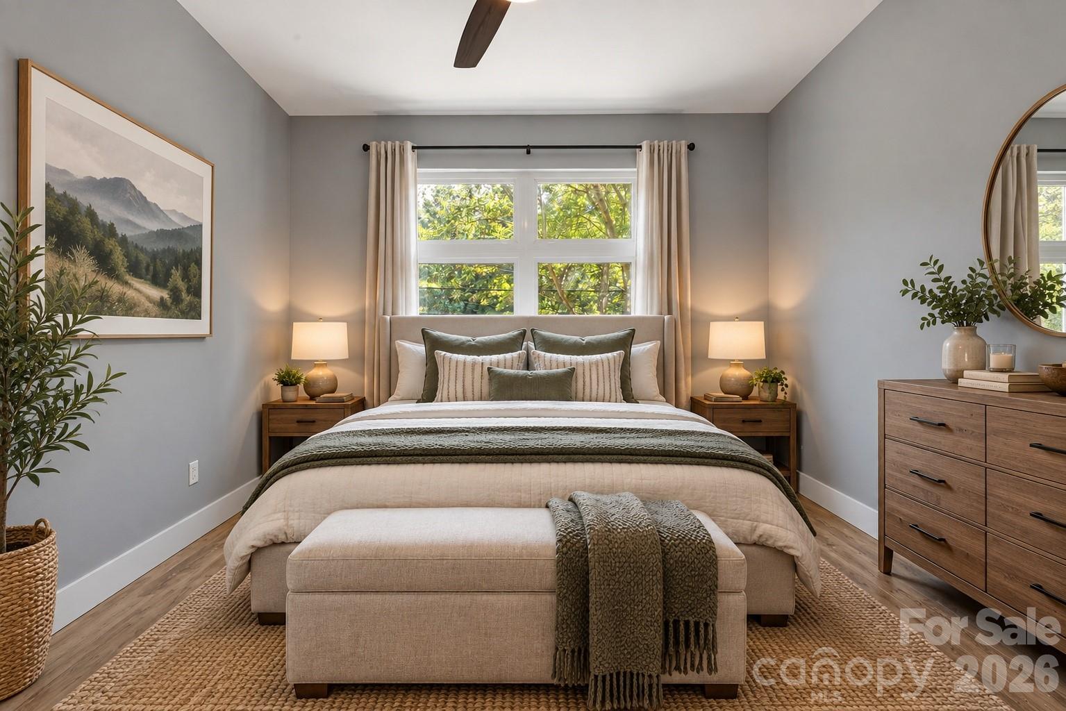Staged bedroom with a layered upholstered bed beneath a large window overlooking leafy trees.