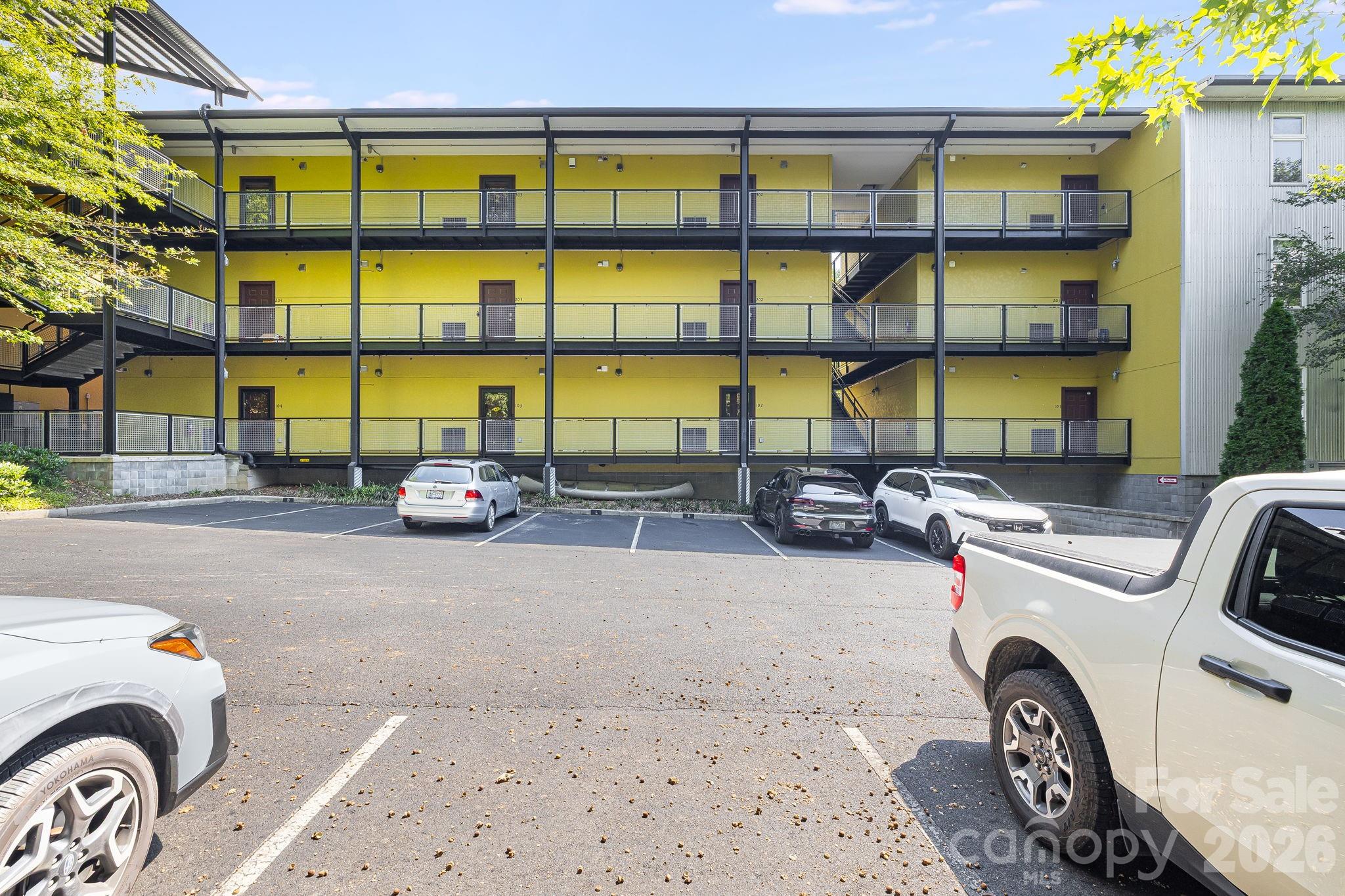 Yellow three-story condominium building with covered balconies and a parking lot in front.