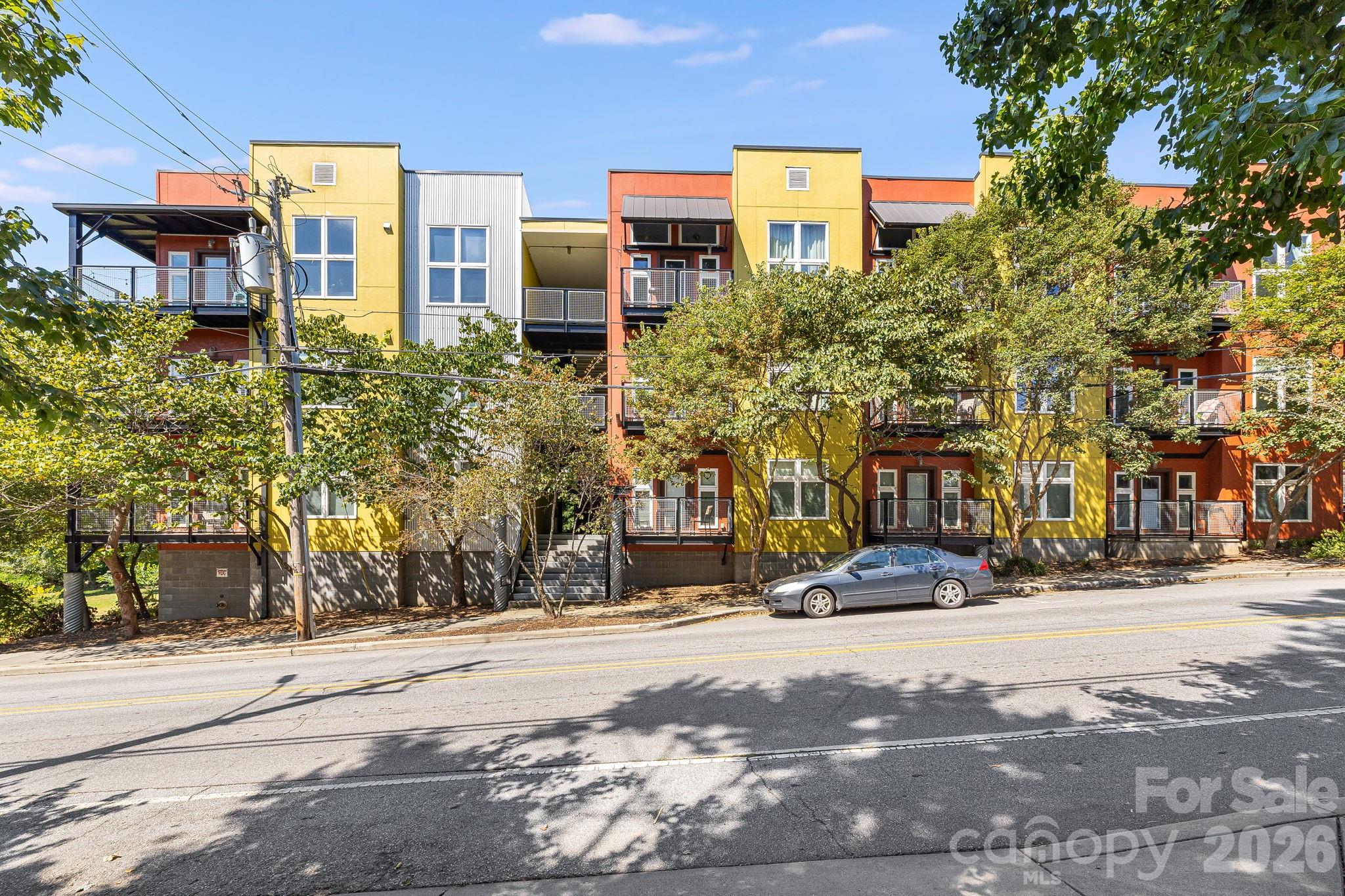 Colorful multistory condominium building with balconies, leafy trees, and a gray car parked out front.