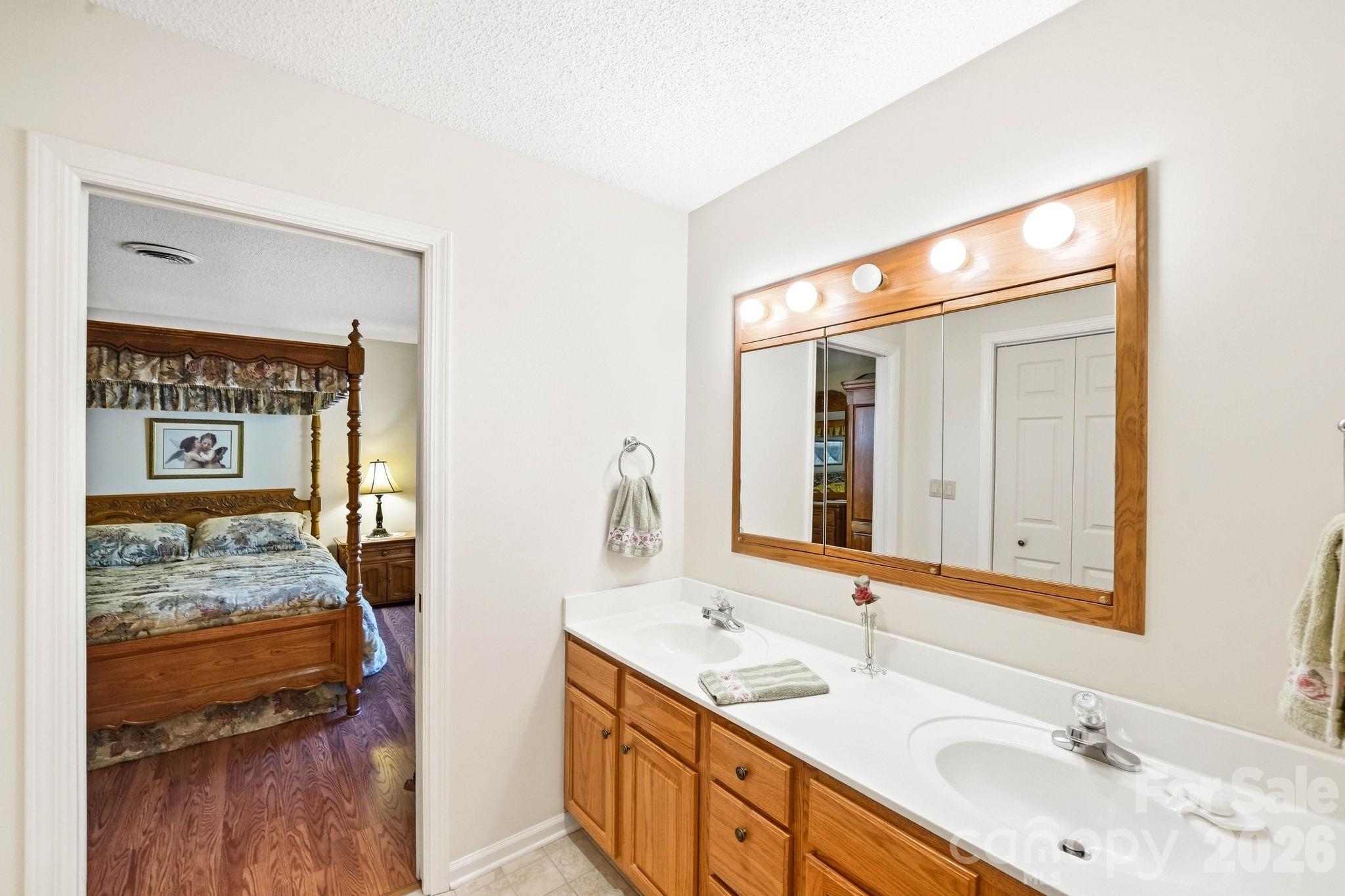 Bathroom with a double-sink wood vanity and lit mirror, opening onto a bedroom with a four-poster bed.