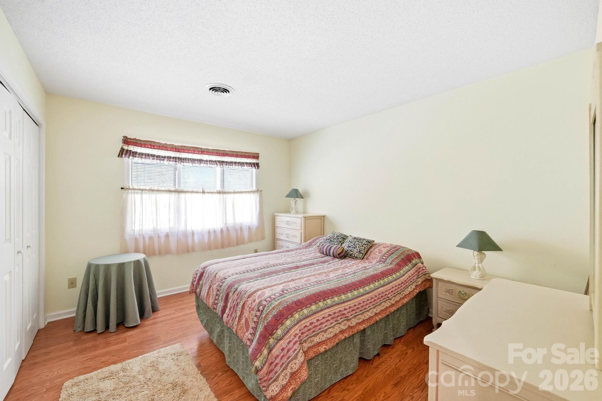 Furnished bedroom with a colorful patterned quilt, hardwood floors, and a curtained window.